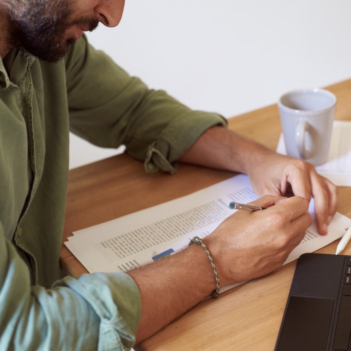 main in green shirt with a beard, writing on a piece of paper with a coffee cup next to him